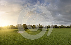 Golf course with lawn. Evening landscape in the park. Blue sky with clouds and bright sunlight with rays at sunset