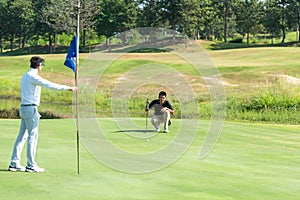 Golfer asian man with friend playing aiming shot for putting ball on the hole with club on green course.