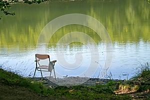 Gone fishing, perhaps, chair left by lake edge. Summer. Background, nobody there in lakeside seat.