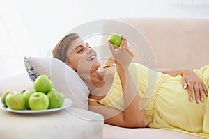 Good for the baby. A happy pregnant woman relaxing on her couch and enjoying an apple.