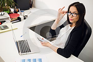Good business talk. Cheerful young beautiful woman in glasses talking on mobile phone and using laptop with smile while