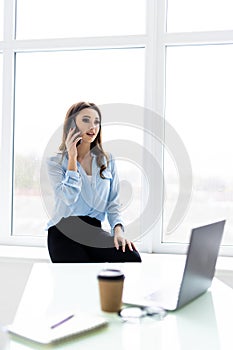 Good business talk. Cheerful young beautiful woman in glasses talking on mobile phone and using laptop with smile while sitting at