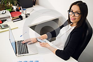 Good business talk. Cheerful young beautiful woman in glasses talking on mobile phone and using laptop with smile while