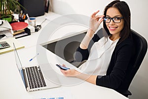 Good business talk. Cheerful young beautiful woman in glasses talking on mobile phone and using laptop with smile while