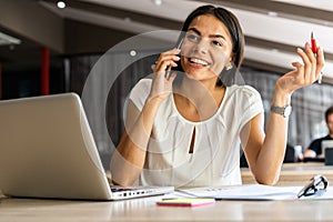 Good business talk. Cheerful young beautiful woman talking on mobile phone and using laptop with smile while sitting at