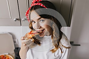 Good-humoured girl eating pizza in kitchen. Close-up shot of positive woman enjoying her dinner.