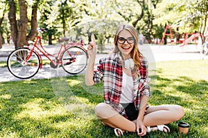 Good-humoured young woman sitting on the grass with sincere smile. Outdoor photo of blithesome cauc