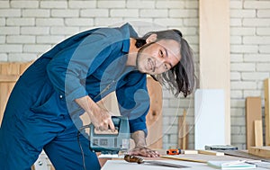 Good looking Asian carpenter wearing coverall dress working for DIY jobs in carpenter room with several kinds of woods and types