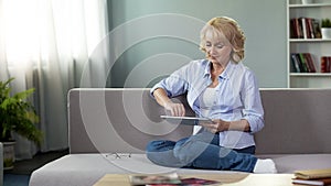 Good-looking blonde senior woman sitting on sofa and scrolling on tablet, apps