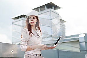 Good looking businesswoman in beige suit, brown trousers and helmet sit on the roof and hold laptop