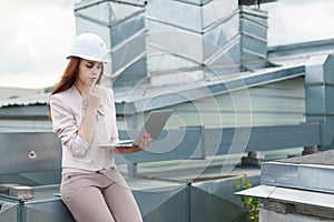 Good looking businesswoman in beige suit, brown trousers and helmet sit on the roof and hold laptop