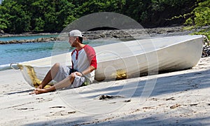 Good looking castaway man sitting in the beach by a wrecked boat waiting for help with ocean and jungle in the background.