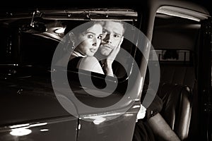 Good looking couple, handsome man in suit, beatiful woman in red dress, are discovered embracing in vintage car