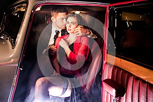Good looking couple, handsome man in suit, beatiful woman in red dress, embrace passionately in vintage car