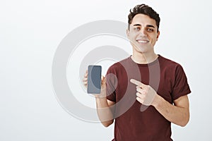 Good-looking friendly male shop assistant in casual red t-shirt, showing brand new smartphone and pointing at device