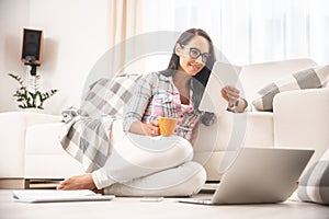 Good looking girl in glasses reading a tablet and drinking coffee leaned against a couch and sitting on a floor