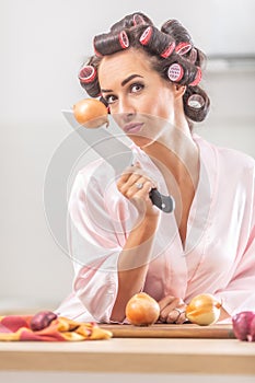 Good-looking girl looks at the camera with an onion stuck on the knife in her hand while standing in night dress in the kitchen