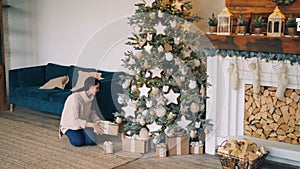 Good-looking girl in warm sweater is bringing gift boxes to Christmas tree, putting them under fir-tree and smiling then
