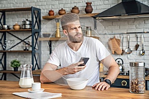 Good looking handsome bearded man is sitting in the kitchen having breakfast cereals and milk, coffee and bananas. holding his