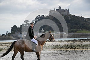 Good Looking Male Horse Rider riding horse on beach in traditional riding clothing with St Michael`s Mount in background