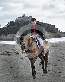 Good Looking Male Horse Rider riding horse on beach in traditional riding clothing with St Michael`s Mount in background