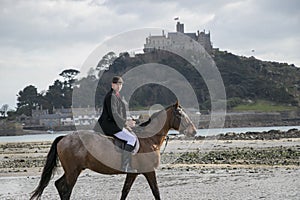 Good Looking Male Horse Rider riding horse on beach in traditional riding clothing with St Michael`s Mount in background