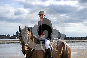 Good Looking Male Horse Rider riding horse on beach in traditional riding clothing with St Michael`s Mount in background