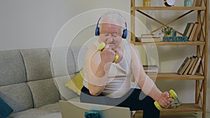 Good looking old man at home in living room sitting on the sofa and training the biceps with weights while listening