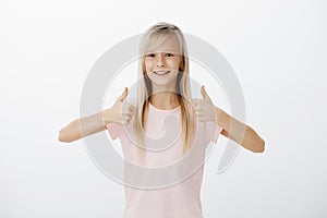 Good-looking positive european little girl with natural fair hair, showing thumbs up and smiling broadly, approving idea