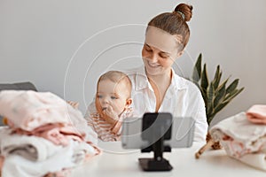 Good looking pretty young female blogger, with hair bun broadcasting livestream at home together with her infant daughter, looking