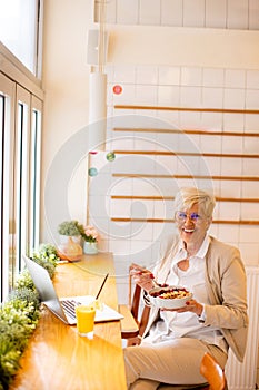 Good looking senior woman working on laptop, drinking orange juice and having a healthy breakfast in cafeteria