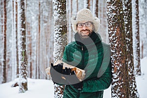 Good looking smiling male wears warm winter clothes holds firewood, stands near tree, spends free time with friends in winter fore