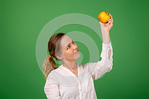 A good-looking woman of forty in a white shirt on a green background smiles and holds a fresh apple in her hands. Healthy eating,