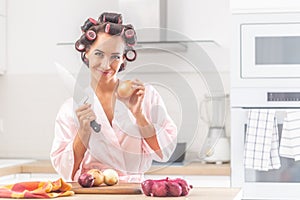 Good looking woman stands in the kitchen holding knife and an onion, wearing hair curlers and night gown