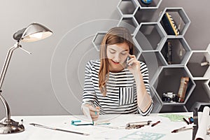 Good-looking young entrepreneur designer with dark hair in striped shirt talking on phone with customer discussing