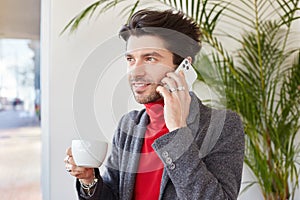 Good looking young glad unshaved brunette male keeping cup of coffee in raised hand and smiling pleasantly, making call while