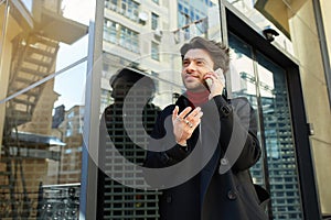 Good looking young handsome bearded brunette male in elegant clothes keeping mobile phone in raised hand while having pleasant