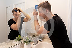 Good Looking Young Man Combing and Looking at His Hair, Beard, and Face in His Home Bathroom Mirror in the Morning Getting Ready f