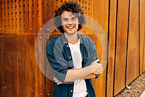 Good-looking young man with curly hair, smiling and standing at modern metal building wall outdoors. Portrait of happy smart