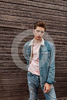 Good-looking young man with hairstyle with stylish blue denim jacket in pink fashionable t-shirt poses near old wooden brown wall
