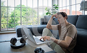 A good-looking young man sitting on a laptop or notebook with a worried, worried and bored expression on his face . A young man