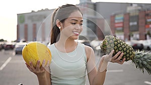 Good-looking young woman holding in her both hands melon and peanapple and weighting what fruit is heavier