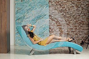 Good-looking young woman make-up and long hair wearing yellow dress posing sitting on blue trendy comfortable sofa