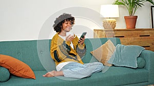 Good morning, African girl drinking coffee holding smartphone sitting on couch at home Woman with cell phone surfing