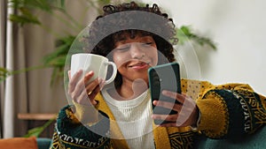 Good morning, African girl drinking coffee holding smartphone sitting on couch at home Woman with cell phone surfing