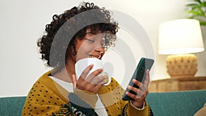 Good morning, African girl drinking coffee holding smartphone sitting on couch at home Woman with cell phone surfing