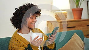 Good morning, African girl drinking coffee holding smartphone sitting on couch at home Woman with cell phone surfing