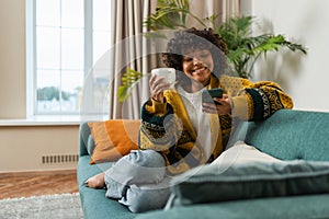 Good morning, African girl drinking coffee holding smartphone sitting on couch at home Woman with cell phone surfing