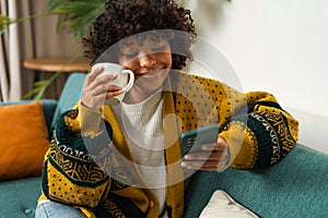 Good morning, African girl drinking coffee holding smartphone sitting on couch at home Woman with cell phone surfing