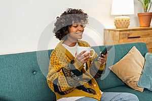 Good morning, African girl drinking coffee holding smartphone sitting on couch at home Woman with cell phone surfing